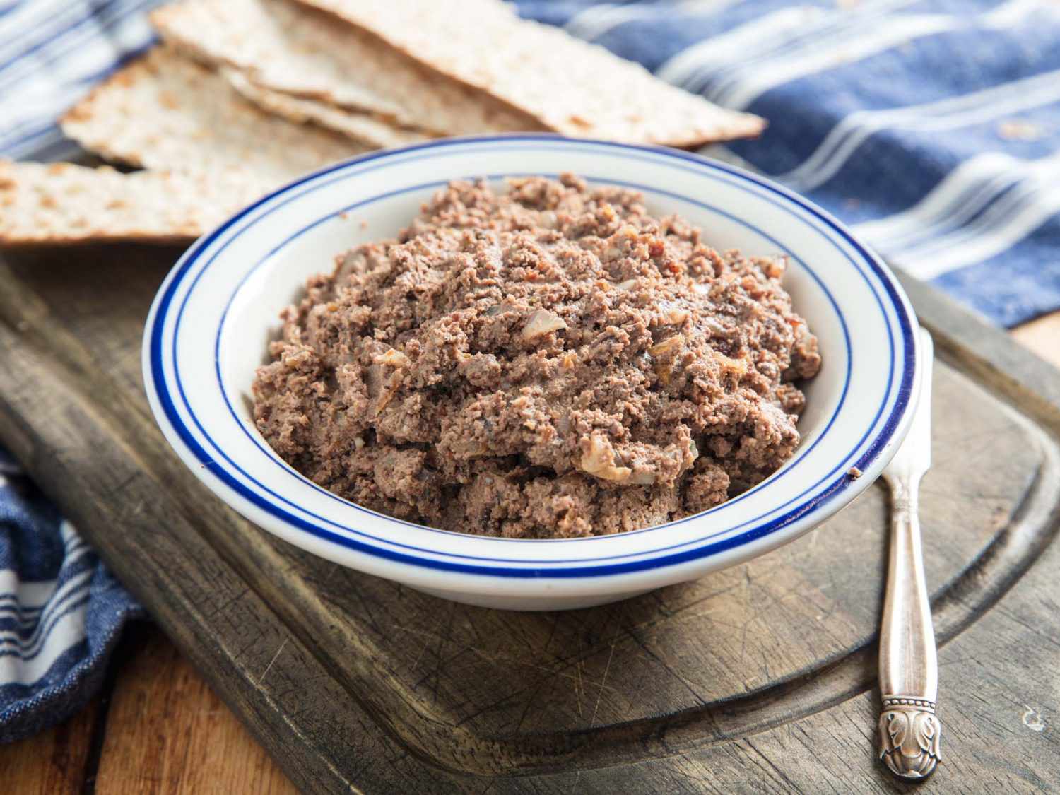 一个博wl of chopped liver on a wooden board with matzo and a knife.