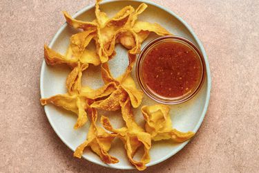 Golden, fried crab rangoons on a blue ceramic plate which also holds a small glass bowl of sauce, on a pink stone background.