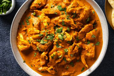 Chicken tikka masala in a white speckled ceramic bowl. In the top left corner is a small bowl holding cilantro leaves and on the top right edge of the image is a plate holding naan bread.