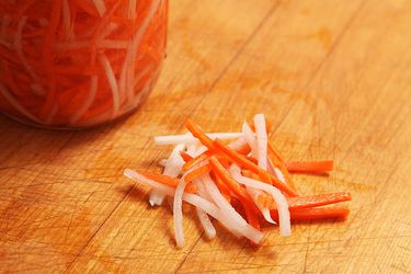 一罐越南腌制萝卜和胡萝卜，用来装饰越南越南三明治，一小堆腌制蔬菜放在坛子前面的木柜台上。