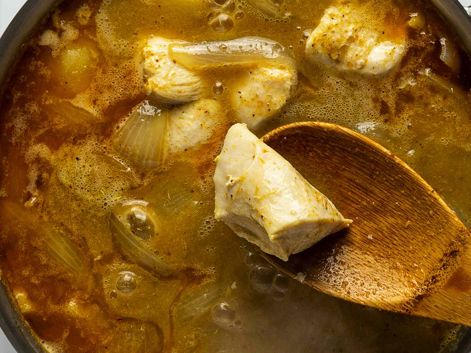 A close-up shot of the contents of the saucepan after the chicken has been added. A wooden spoon is lifting a piece of chicken out of the pan to demonstrate its cooked color and texture.