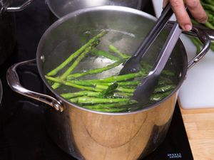 Asparagus being blanched in a large stock pot filled with water. Someone is removing the asparagus with a pair of tongs.