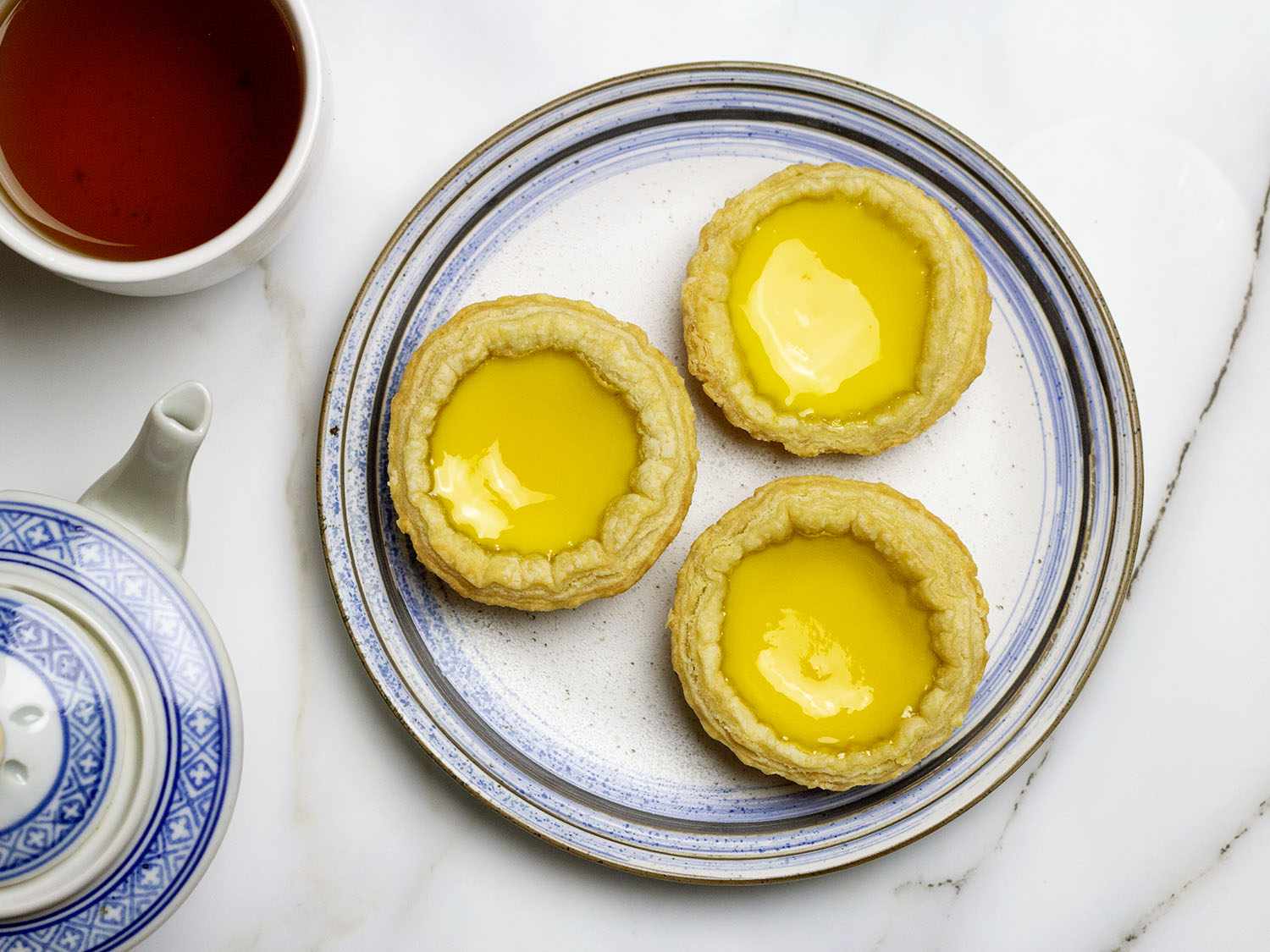 Three Hong Kong egg custard tarts on a plate net to a cup of tea and a tea pot.