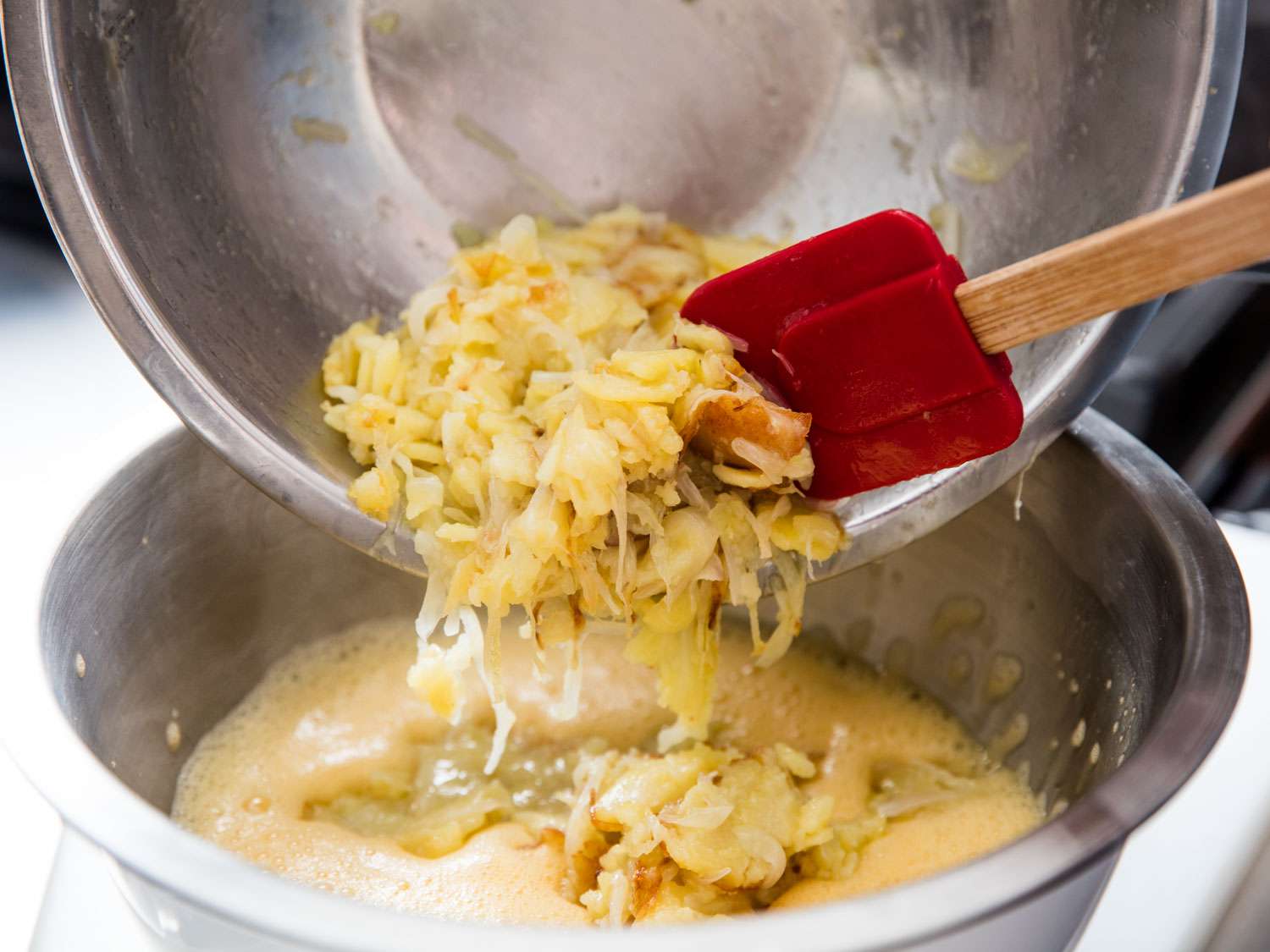 Pouring cooked potatoes and onions into bowl of frothed eggs.