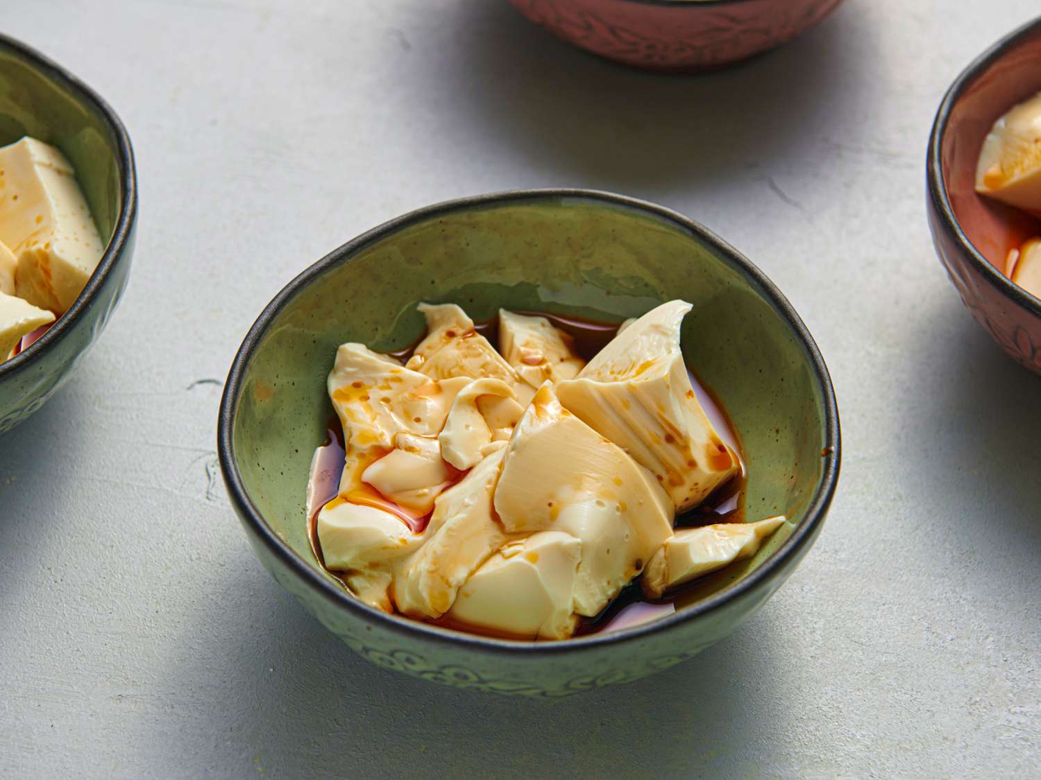 A small, irregularly round glazed ceramic bowl holding soft peaks of silken tofu covered with chili oil, soy sauce, and sesame oil.