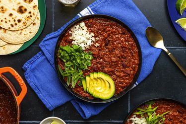 辣椒顶部有一大碗素食beansliced avocado, scallions, cilantro, and white onion. The periphery of the image contains a wide variety of plates and bowls holding tortillas, sliced limes, and additional chili.