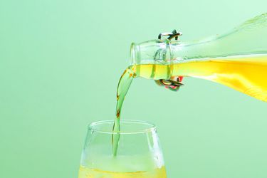 A glass bottle containing elderflower cordial, pouring the liquid into a glass containing ice.