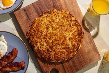 A deeply browned rÃ¶sti sits on a wooden cutting board, with plates of fried eggs and bacon, and glasses of orange juice around it on the table.