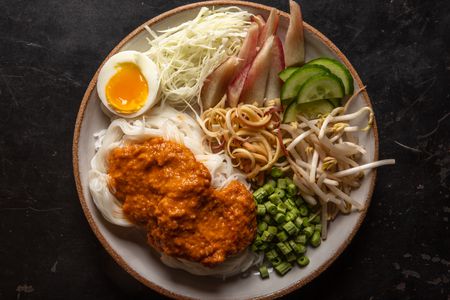 Overhead of a serving plate of noodles dressed with fish curry, with assorted vegetable garnishes and a soft-cooked egg on the side.