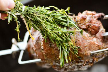 Boneless leg of lamb on a rotisserie, being basted with a bundle of fresh herbs.