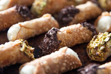 Close up of cannolis lined up in rows. Some of them have had their ends dipped in shaved chocolate, others have been dipped in chopped pistachios.