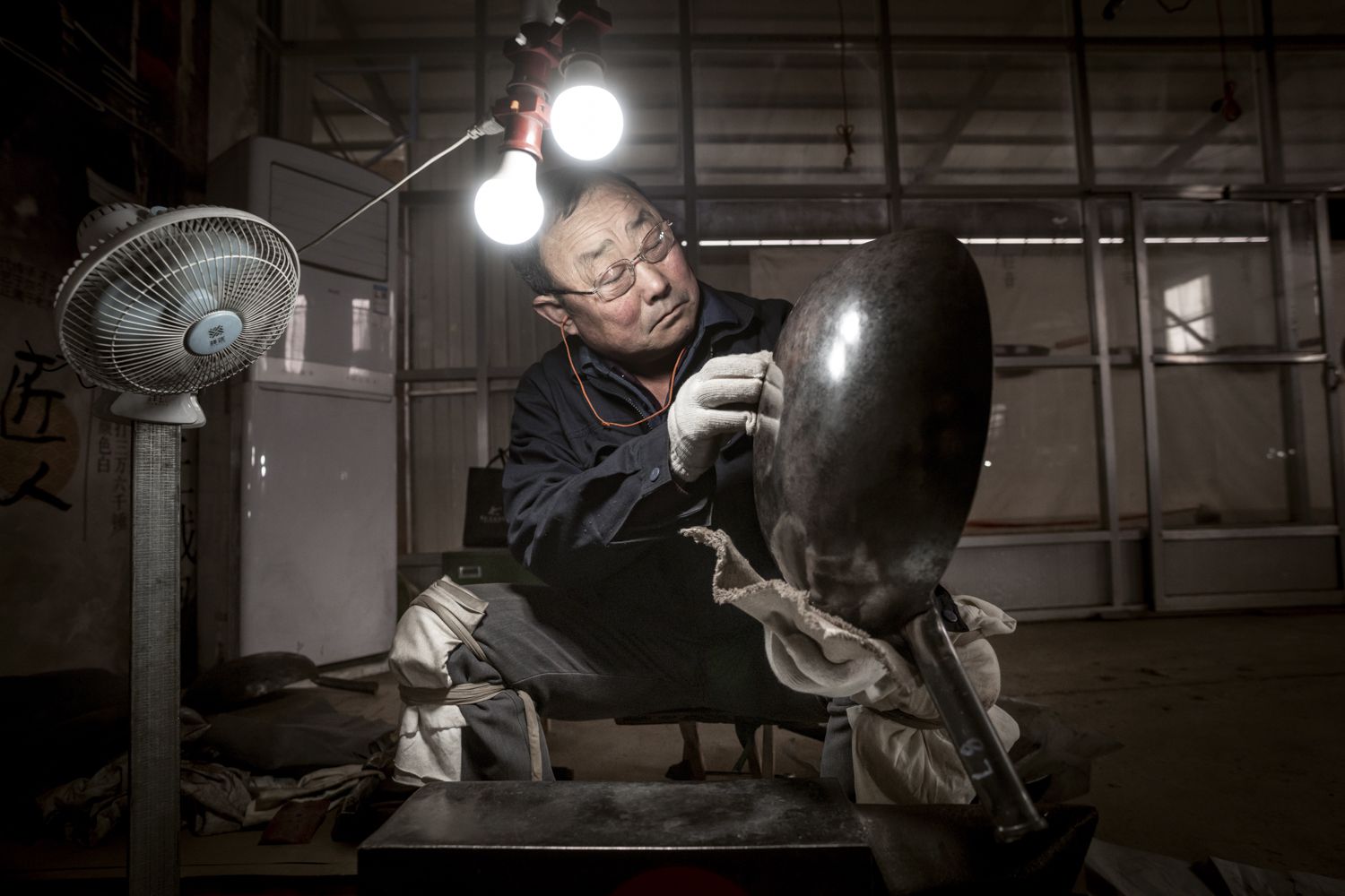 A factory worker polishes a hand-hammered wok under two lightbulbls