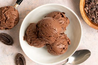 四勺巧克力冰淇淋装在一个圆形的白色陶瓷碗里。碗的外围是一个金属勺子，一个装着可可粒的小木碗，两片巧克力和一个装着冰淇淋的勺子。