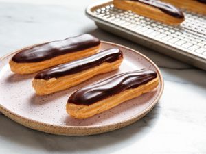 小粉红斑点炻器平台e with three classic éclairs on it, each topped with chocolate icing. A bakers rack with more éclairs is in the background.