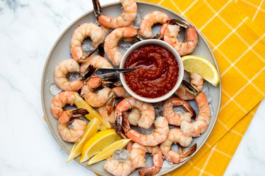 Shrimp cocktail on a ceramic plate with cocktail sauce and sliced lemons, on a stone background with a yellow dish towel.