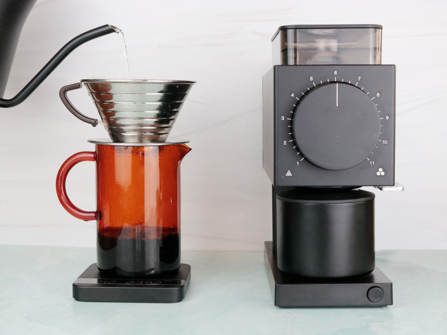 a gooseneck kettle pouring water into a pourover coffee dirpper next to a coffee grinder