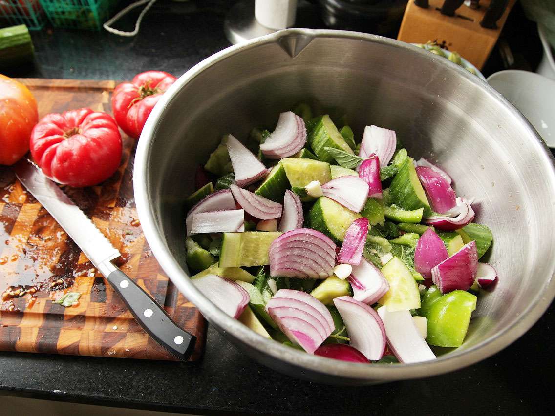 Chunks of red onion and cucumber in a large steel mixing bowl for gazpacho.