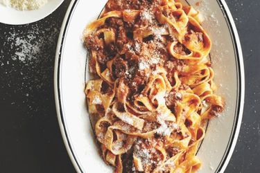 Lamb bolognese tossed with fettucine noodles and sprinkled with freshly grated cheese on a white oval platter.