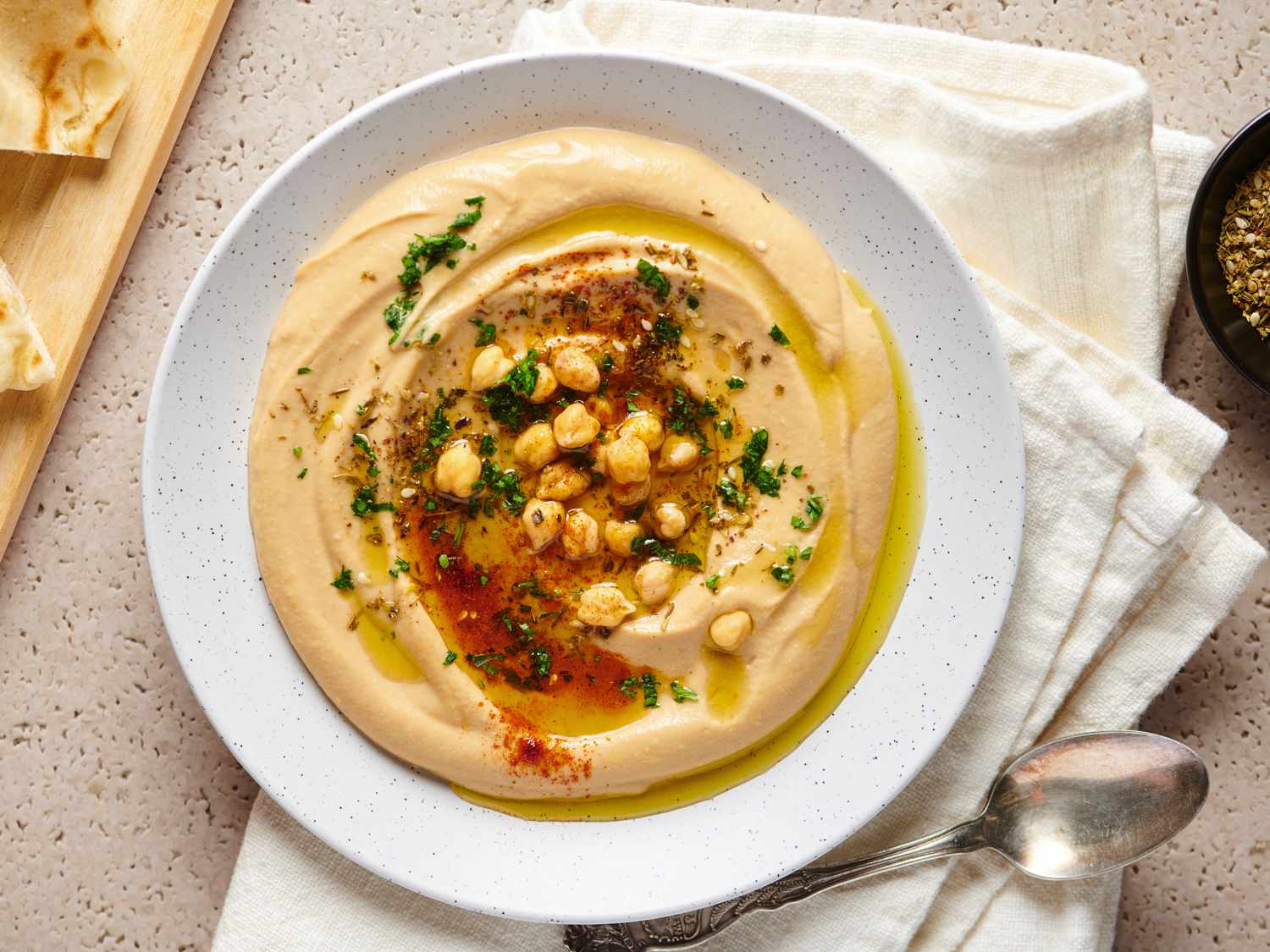 Hummus served on a wide, shallow plate, drizzled with olive oil and sprinkled with zaâatar, paprika, warmed whole chickpeas and chopped parsley.