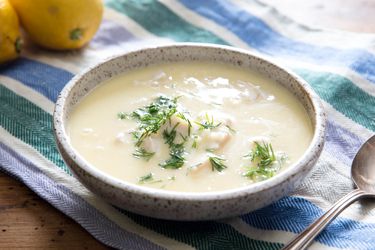 A serving bowl of avgolemono on the table. Some lemons are visible in the background.