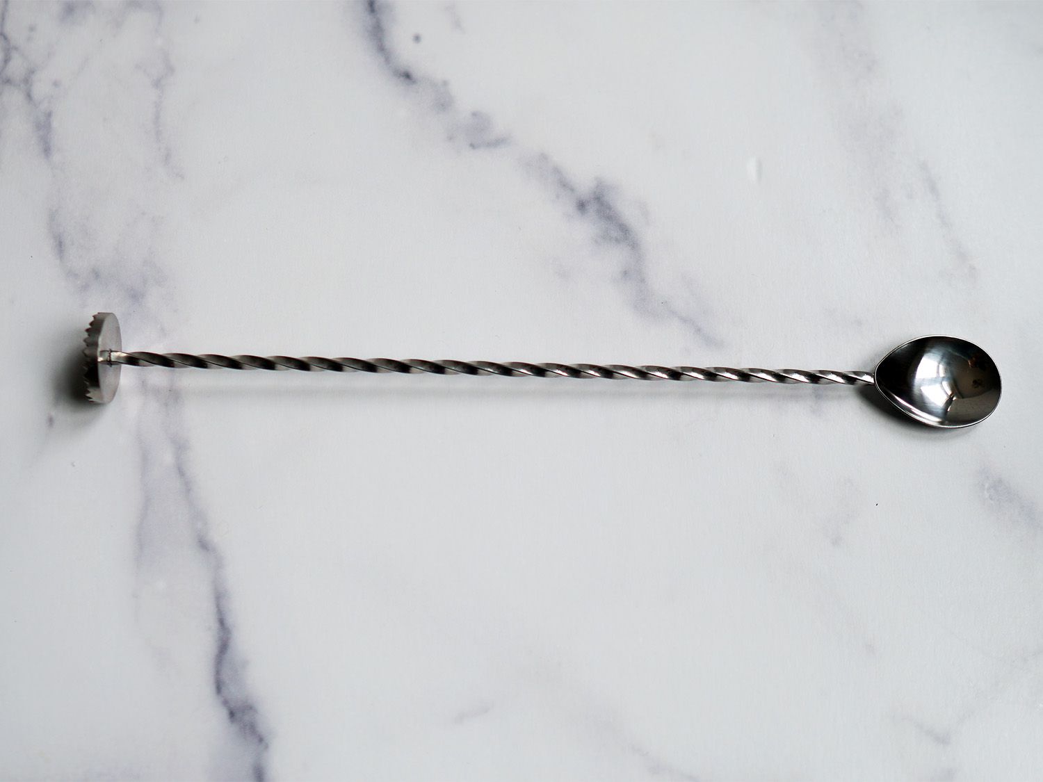 A stainless steel bar spoon with a muddler end on a marble surface