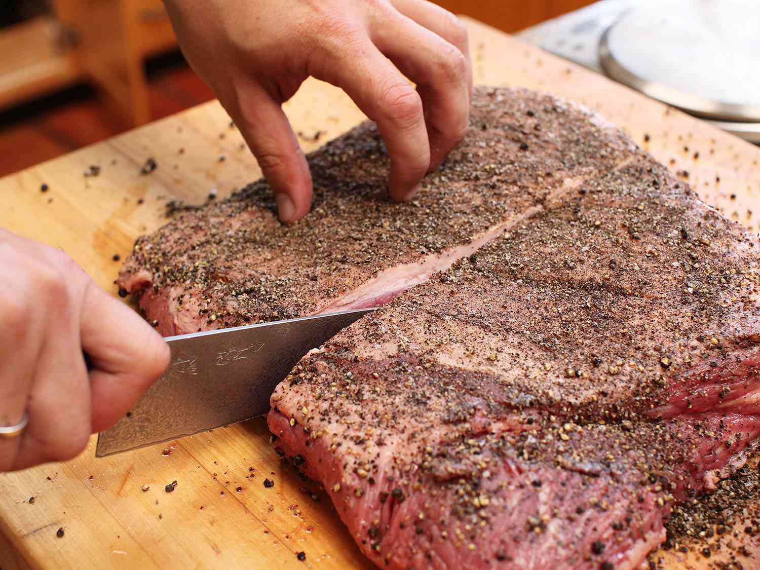 Hands using knife to slice into large piece of peppercorn-crusted raw brisket