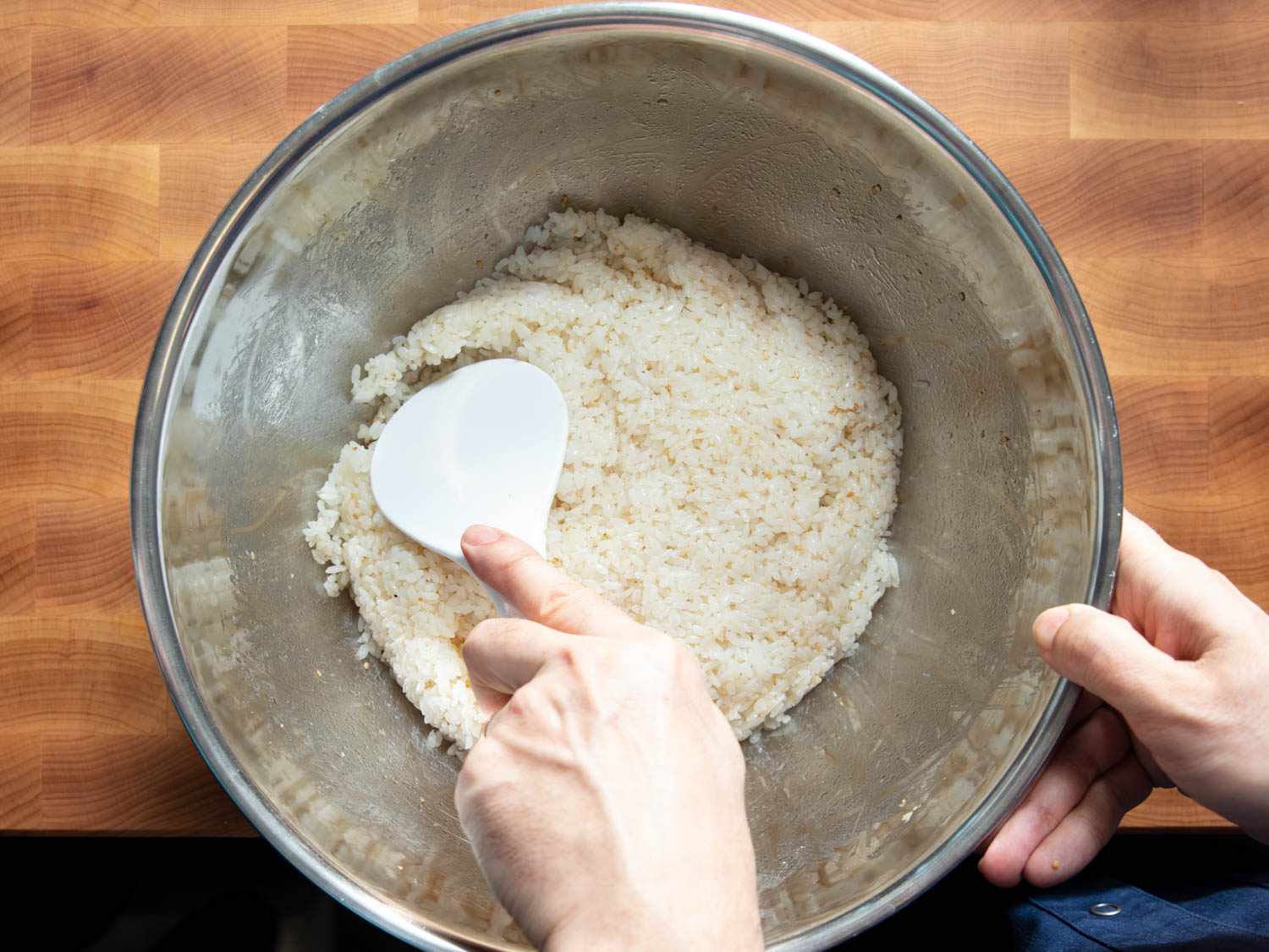 Overhead shot of a person using a white paddle utensil to pat down sticky rice in a mixing bowl.