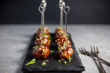 A slate cheese board with eight hoisin-glazed cocktail meatballs on top; each meatball has a metal skewer in it.