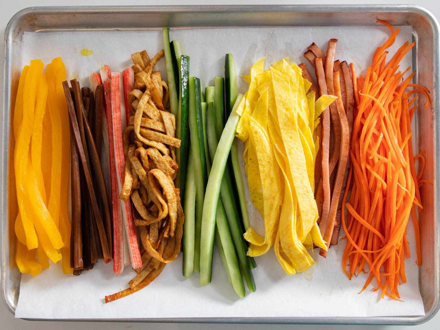 Colorful sliced fillings for kimbap lined up on a baking sheet covered with parchment. The fillings are: pickled radish, braised burdock, imitation crab sticks, cucumber, egg, ham, and carrot.