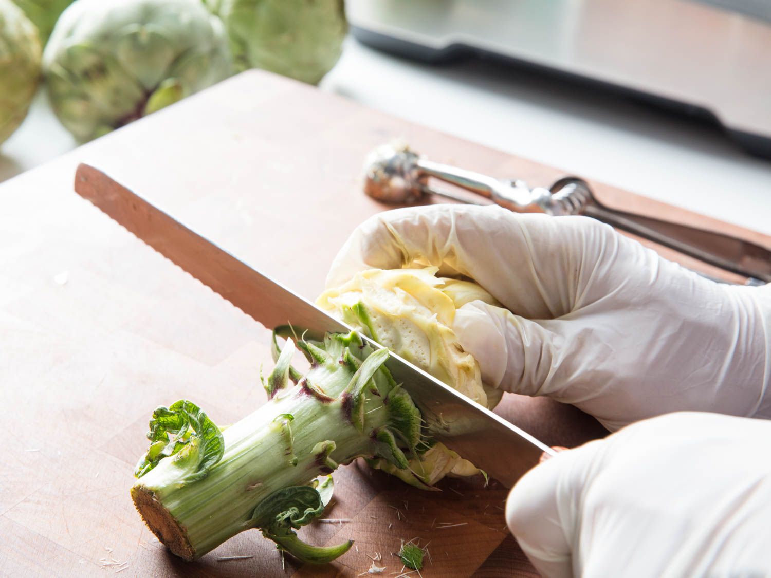 Gloved hands slicing off stem end of an artichoke on a wooden cutting board.