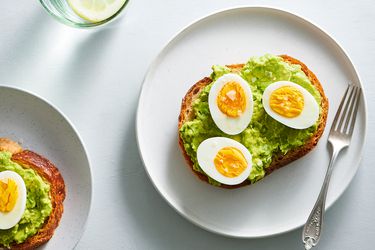 Halved hard-boiled eggs topped with flaky salt and placed on bread topped with mashed avocado. The food is placed on a white ceramic plate with a metal fork.