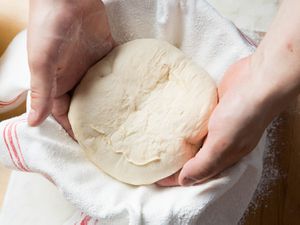 Two hands are placing a ball of bread dough into a bowl lined with a floured kitchen towel.