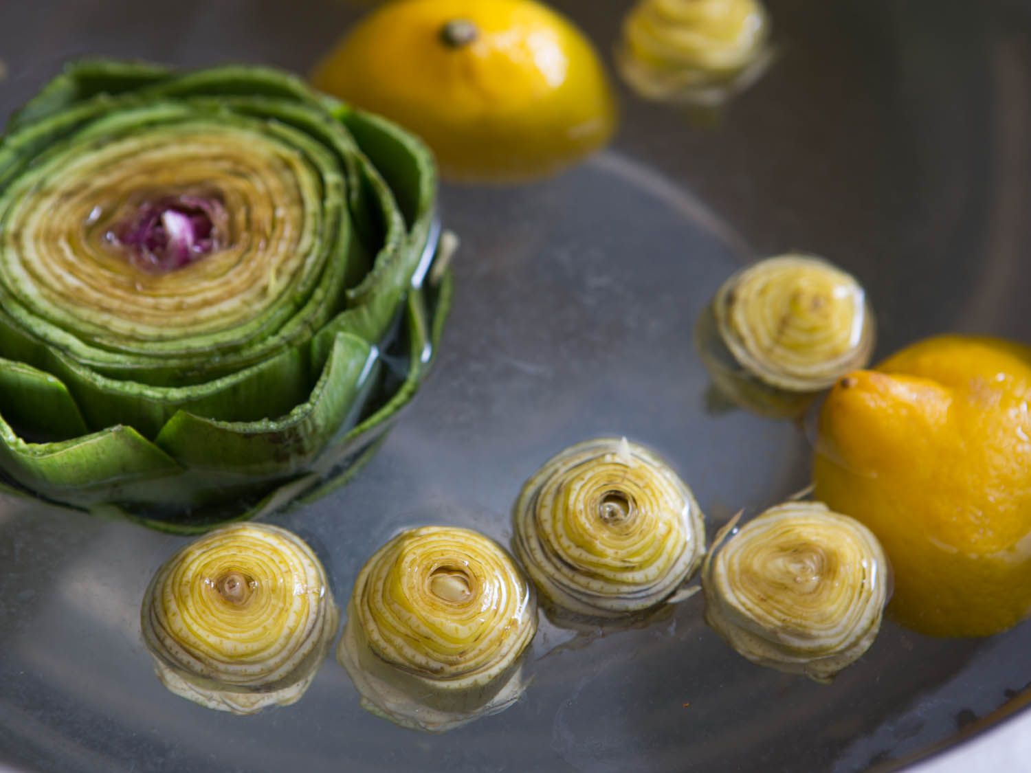 A trimmed large artichoke floating in water with cleaned baby artichokes and lemon halves.