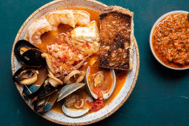 一个serving of cioppino in a bowl, overflowing with plump shrimp, mussels, clams, calamari, fish, and more. There's a piece of deeply toasted sourdough on the side, and a small bowl holding a roasted red pepper condiment.