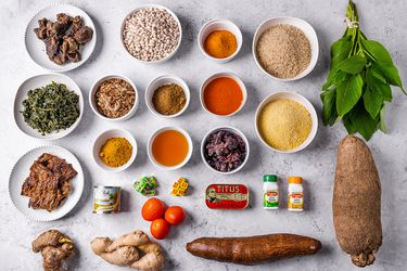 Overhead view of all the essential ingredients needed in a pantry to cook a range of Nigerian dishes. Ingredients range from spices to roots to tinned food and fresh spices, organized in a neat grid