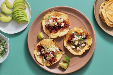 overhead view of three chorizo tacos flanked by plates of sliced avocado and charred corn tortillas