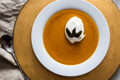 A white bowl of butternut squash soup topped with spiced whipped cream and three fried sage leaves.