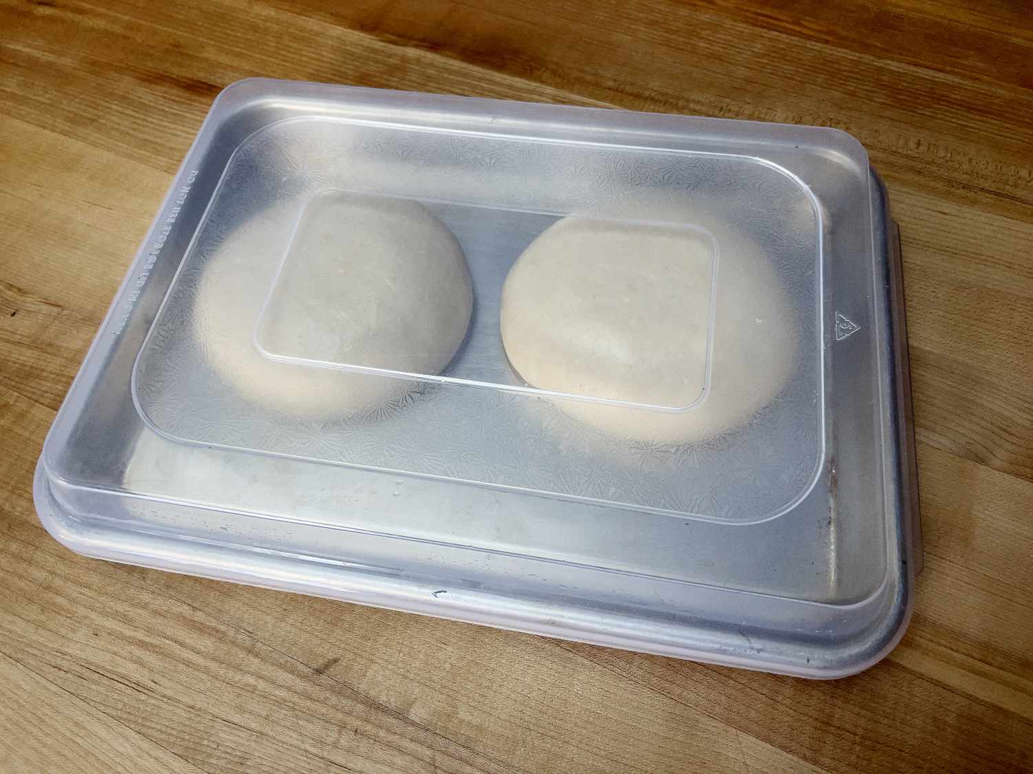 Two balls of dough proofing in a sheet pan with a lid on it