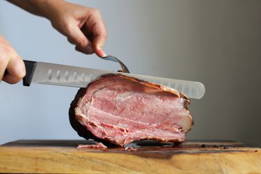 a hand using a slicing knife to cut a piece off of a prime rib roast situated on a wooden cutting board