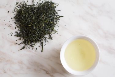 A pile of green tea leaves on a marble countertop, next to a small bowl of brewed green tea.