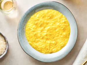 A round ceramic plate holding creamy saffron risotto. There is a glass of liquid in the top left corner and a small bowl of grated cheese on the bottom left edge of the frame.