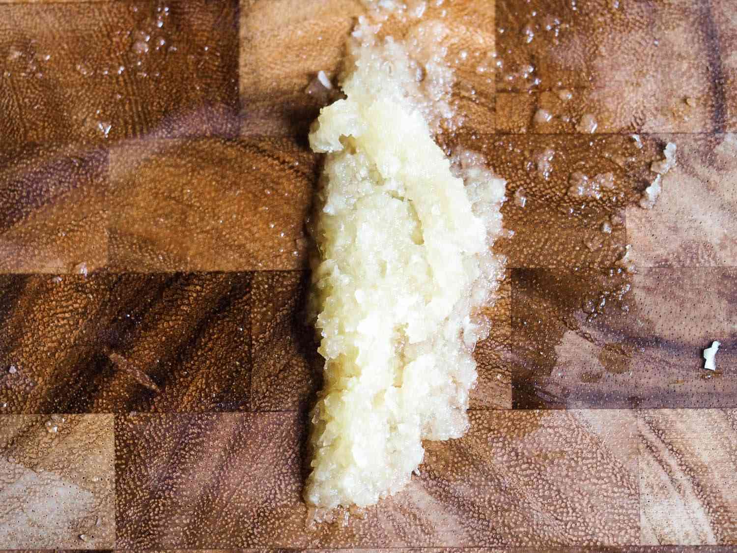 Knife-blade pureed garlic on a cutting board.