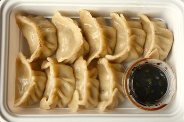 Homemade pork and cabbage dumplings with homemade wrappers, placed in a cardboard take-out container to replicate how they're served in a restaurant. There is a small plastic container of dipping sauce in the bottom right corner of the container.