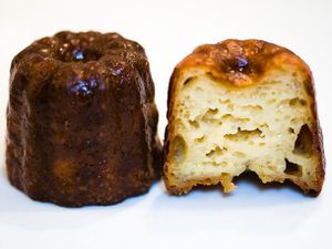 A whole canelé next to another canelé cut open to reveal its custardy interior.