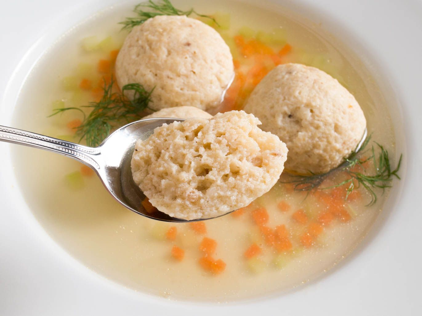 Close-up of a spoon holding a half-eaten matzo ball above a bowl of soup.