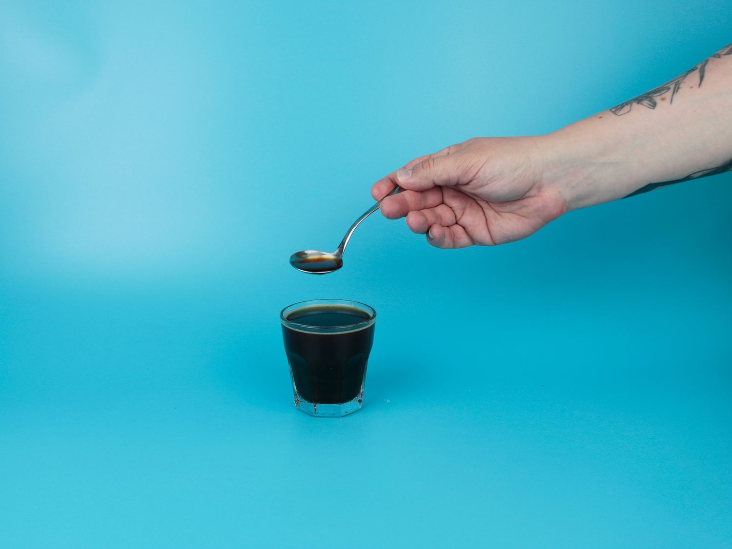 an arm lifting a spoonful of brewed coffee from a glass of water and coffee grounds brewing