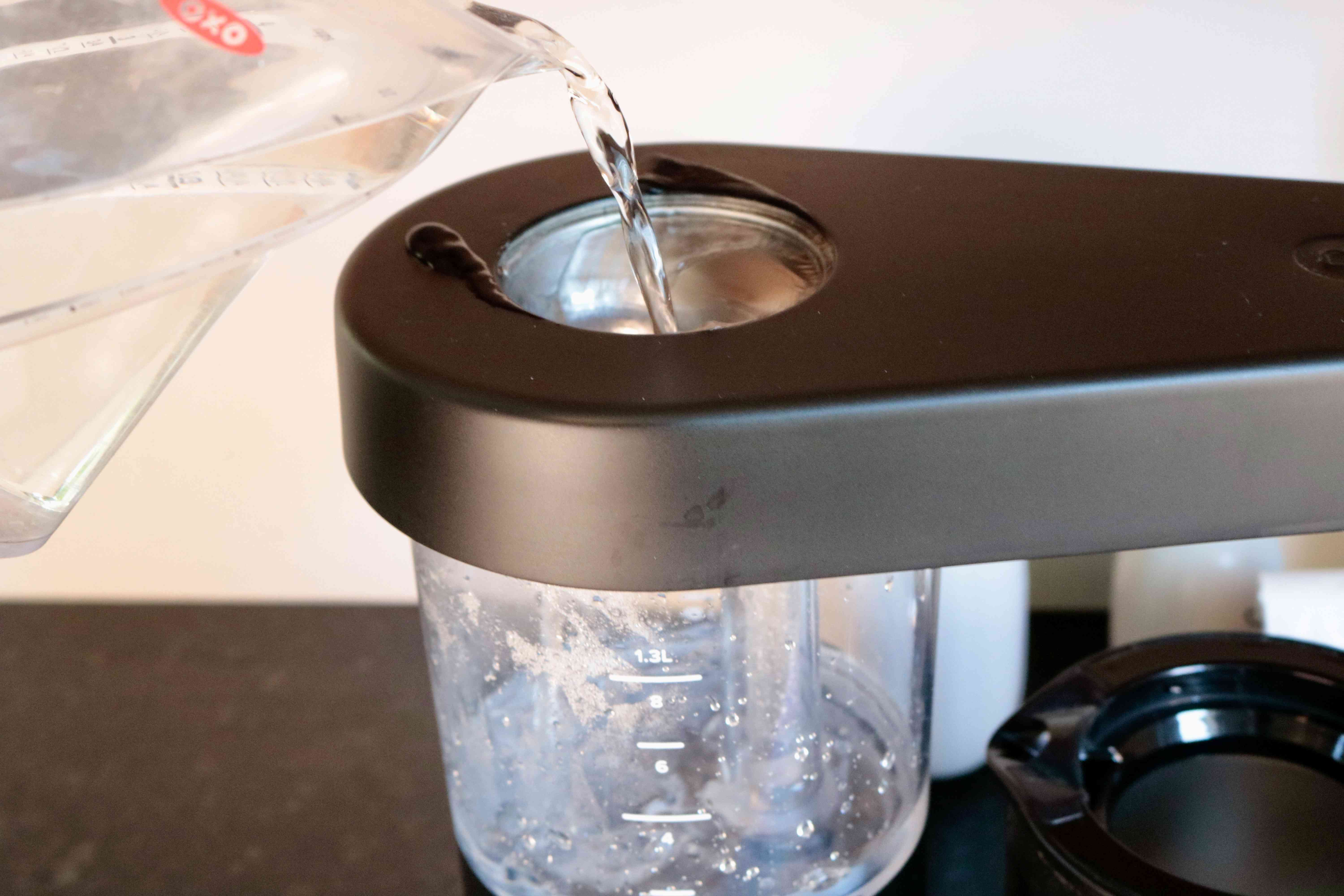 A measuring cup pouring water into a coffee maker's water reservoir