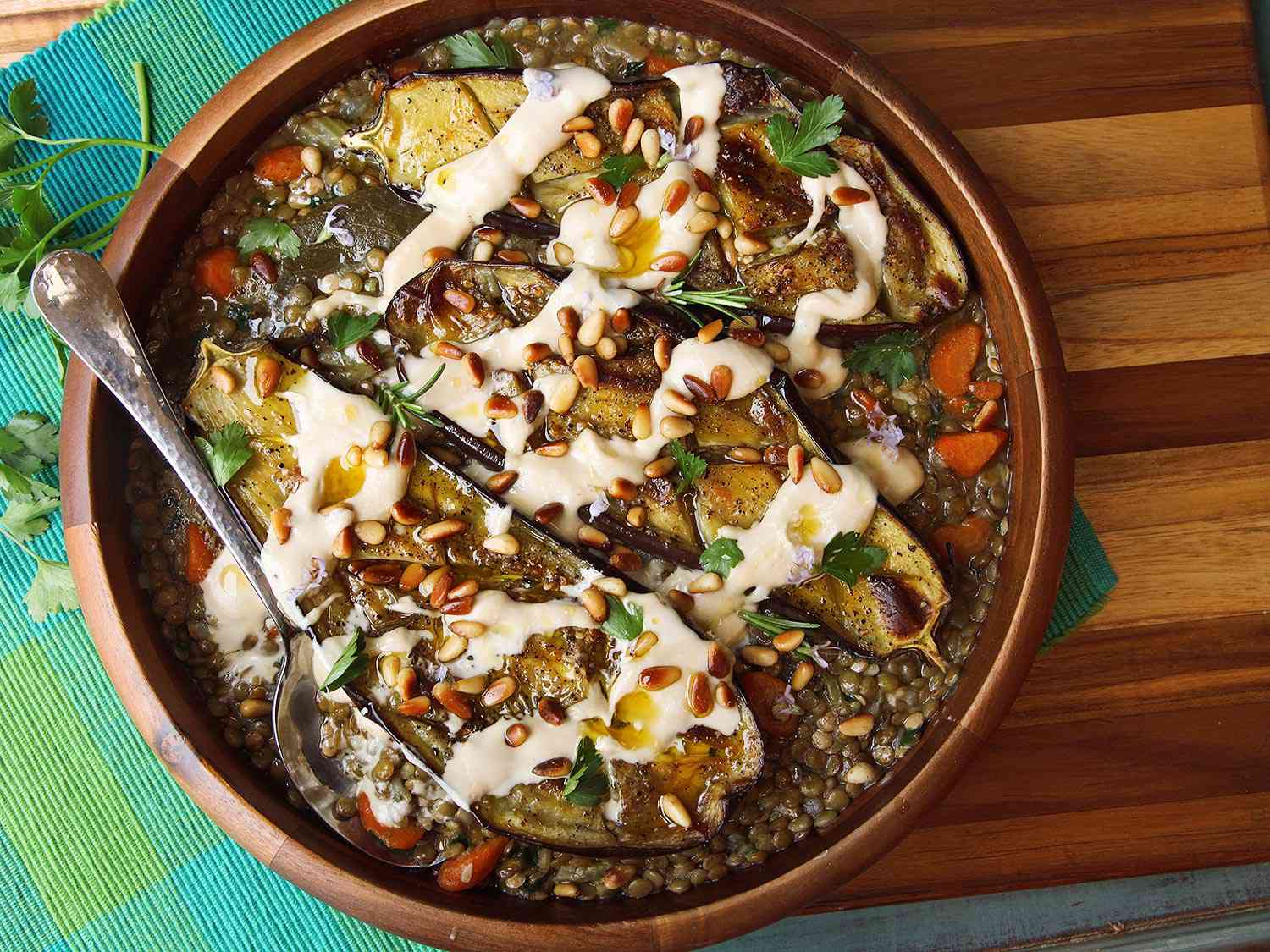 Overhead shot of a serving bowl of roasted eggplant on a bed of lentil stew topped with cream tahini, toasted pine nuts, and chopped herbs