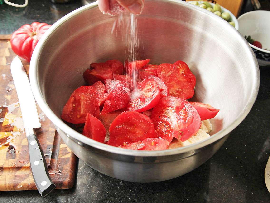 A hand sprinkling salt onto tomatoes in a large steel mixing bowl with bread underneath.
