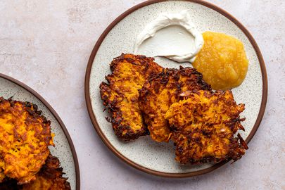 Three crispy sweet potato latkes on a ceramic plate alongside apple sauce and sour cream.